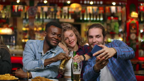 African-american-with-european-friends-sitting-in-a-bar-takes-a-selfie.-A-large-group-of-friends-sits-in-a-barz-at-one-table-chatting-drinking-beer-and-taking-pictures.
